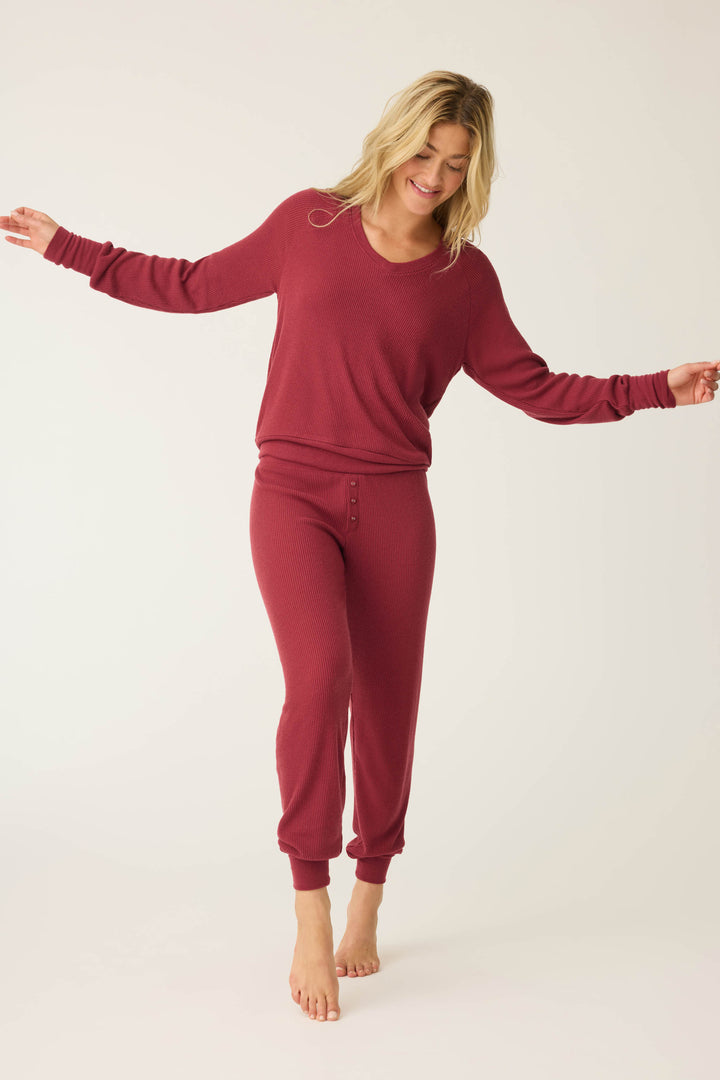 A smiling barefoot woman in the P.J. Salvage Textured Essentials PJ Set—featuring a matching long-sleeve maroon top and jogger-style pants—stands with arms outstretched, looking down against a plain light background.