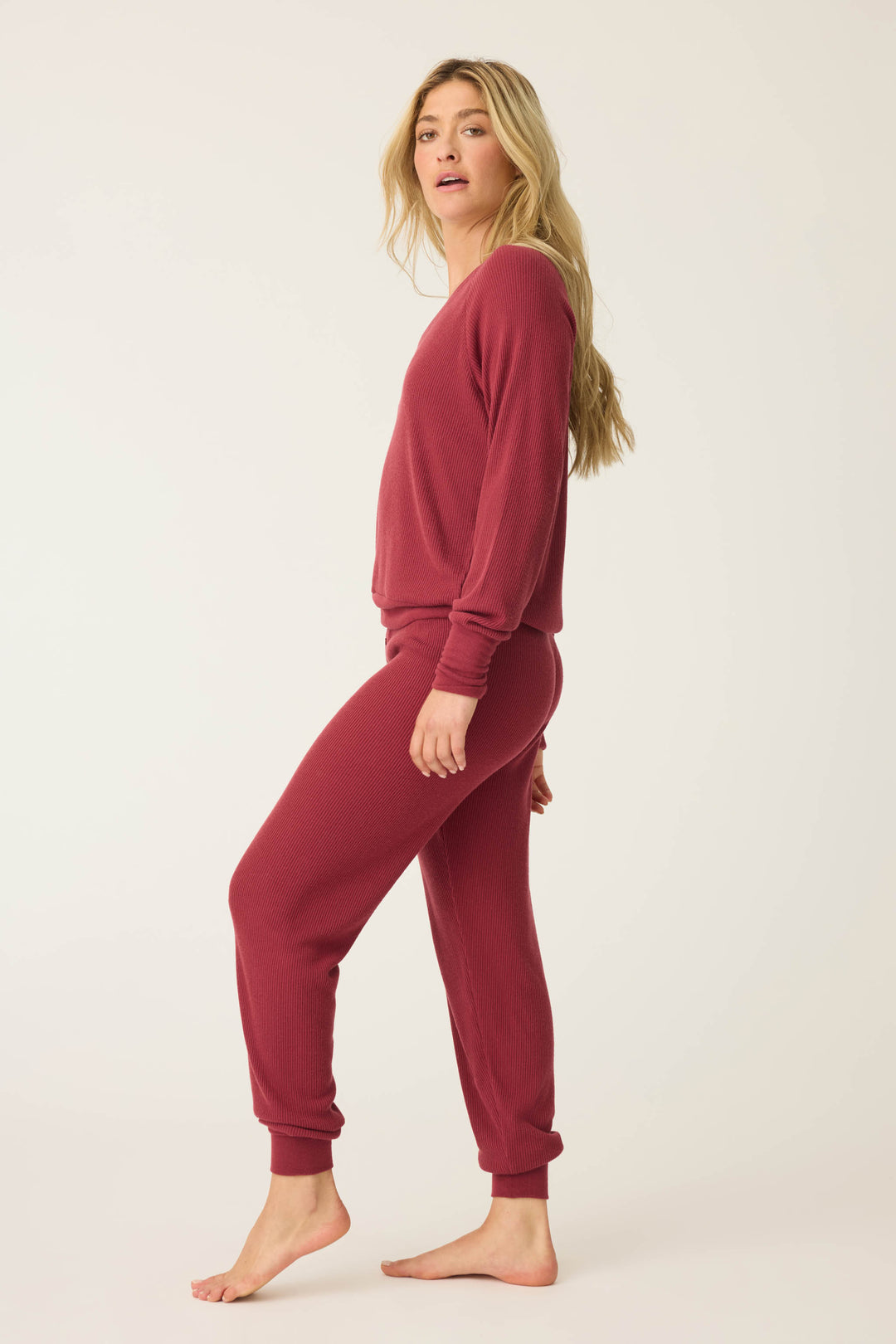 A person with long blonde hair stands barefoot, wearing the P.J. Salvage Textured Essentials PJ Set—a matching maroon long-sleeve top and pants—facing sideways with their head slightly turned toward the camera against a plain light background.