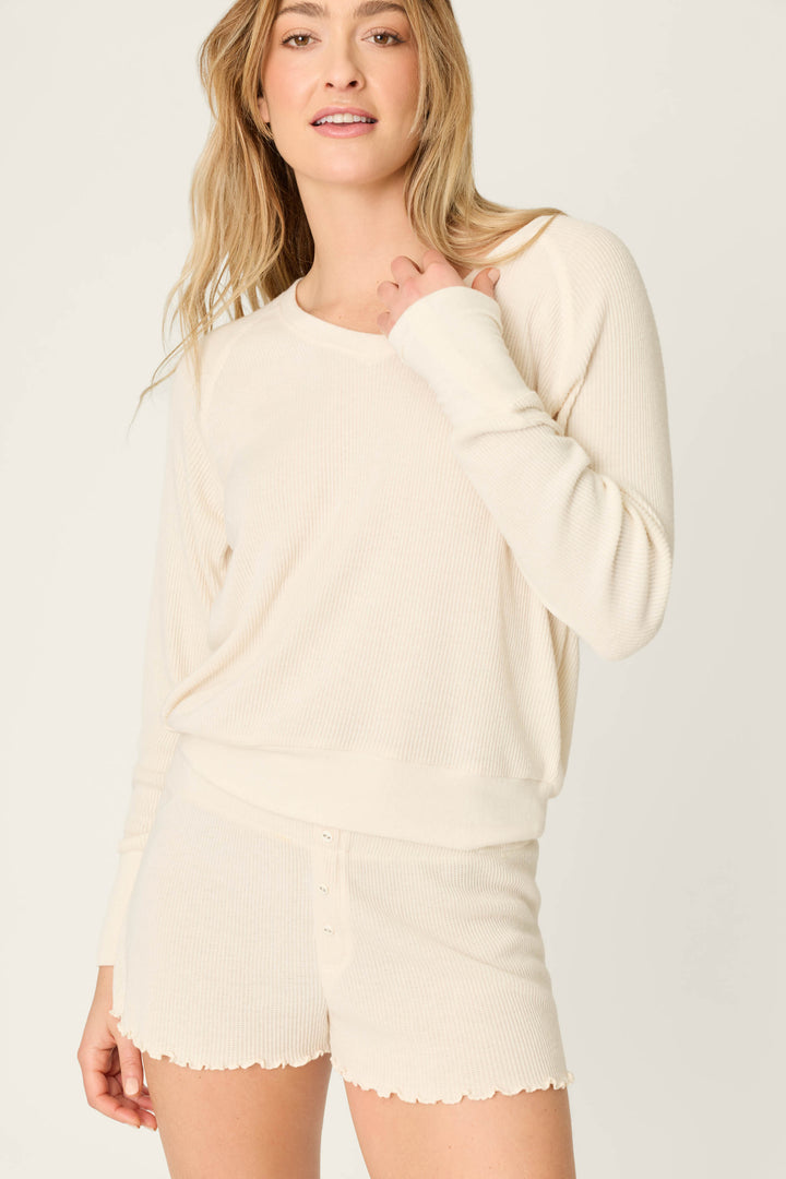 A person with long, wavy blonde hair wears the P.J. Salvage Textured Essentials Stone Short Lounge Set, featuring a ribbed off-white long-sleeve top and matching shorts, standing against a simple light background and looking at the camera.