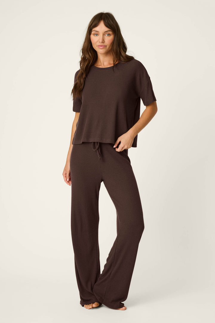 A woman stands barefoot against a plain background, wearing the P.J. Salvage Textured Essentials Chocolate Brown Lounge Set—a loose-fitting ensemble featuring a short-sleeve top and matching wide-leg pants—with a relaxed and casual expression.