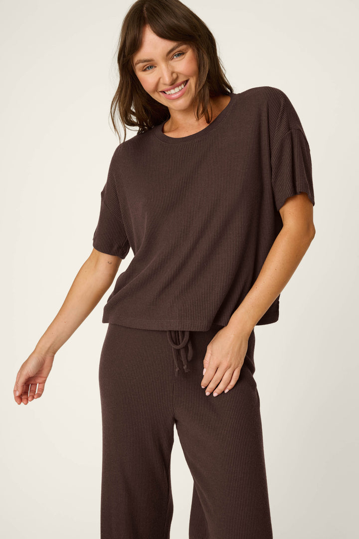 A woman smiles and poses in the P.J. Salvage Textured Essentials Chocolate Brown Lounge Set, featuring a loose-fitting, short-sleeve ribbed top with matching pants, against a plain light background.