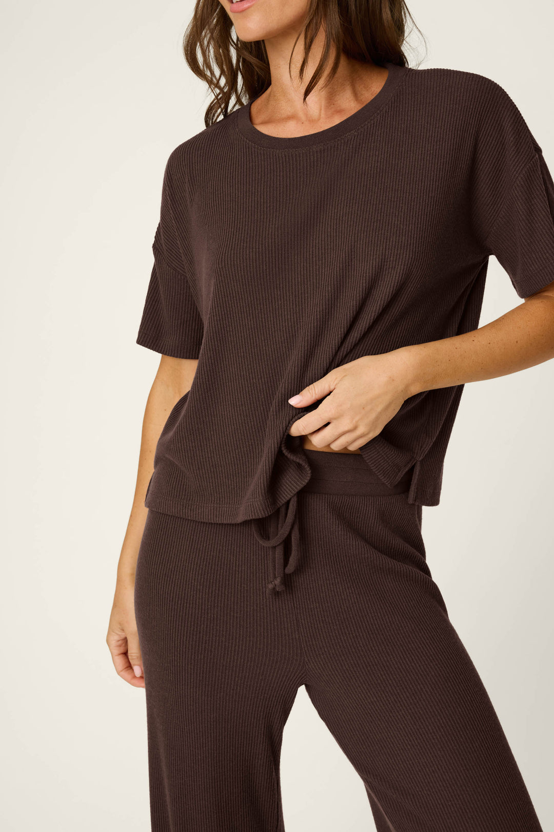 A woman wears the P.J. Salvage Textured Essentials Chocolate Brown Lounge Set, featuring a loose-fitting, ribbed short-sleeve top and matching pants. She slightly lifts the top with one hand, her face partially out of frame against a light background.