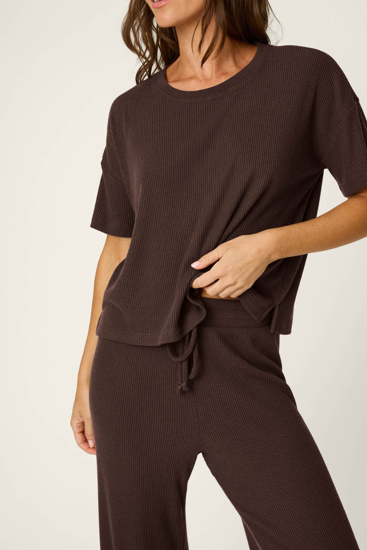A woman wears the P.J. Salvage Textured Essentials Chocolate Brown Lounge Set, featuring a loose-fitting, ribbed short-sleeve top and matching pants. She slightly lifts the top with one hand, her face partially out of frame against a light background.