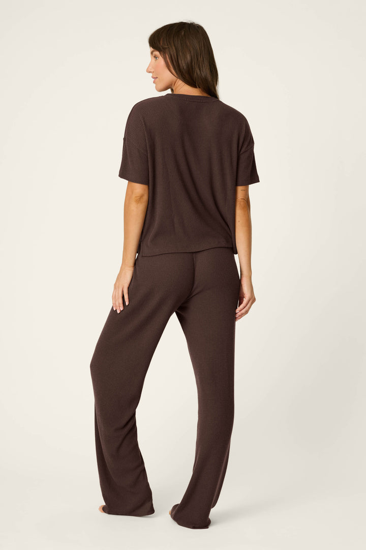 A woman stands barefoot, facing away, wearing the P.J. Salvage Textured Essentials Chocolate Brown Lounge Set—a loose-fitting, short-sleeve top with matching pants—against a plain light background.
