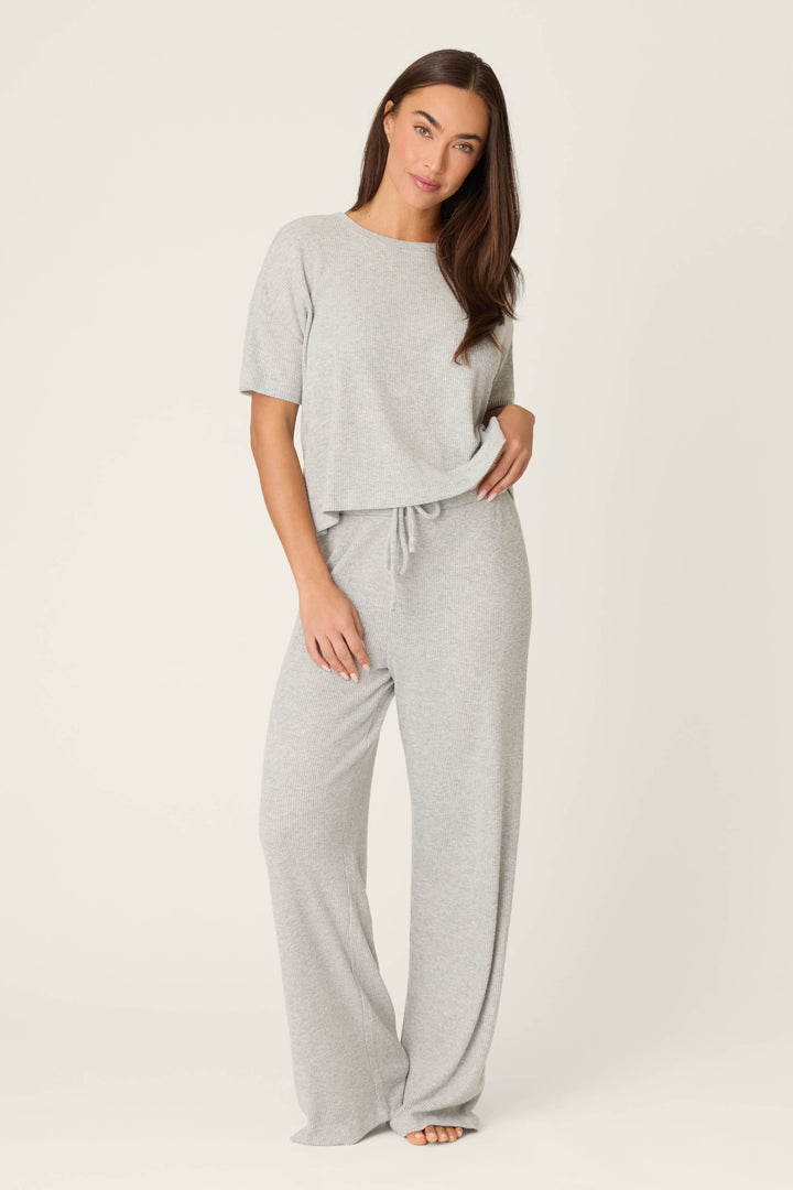 A woman with long brown hair stands barefoot against a plain background, wearing the P.J. Salvage Textured Essentials Heather Grey Long PJ Set featuring a loose short-sleeve top and matching wide-leg drawstring pants.
