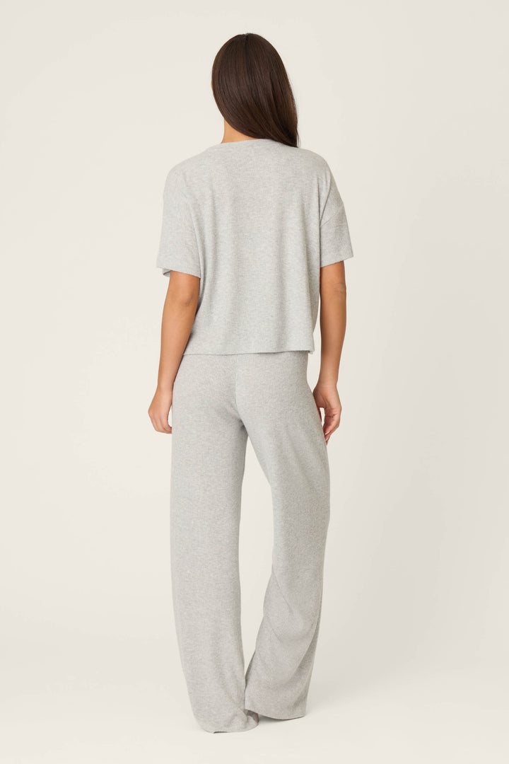 A person with long brown hair stands facing away, wearing the P.J. Salvage Textured Essentials Heather Grey Long PJ Set in an off-white setting. The set features a relaxed fit, short sleeves, and wide-leg pants.