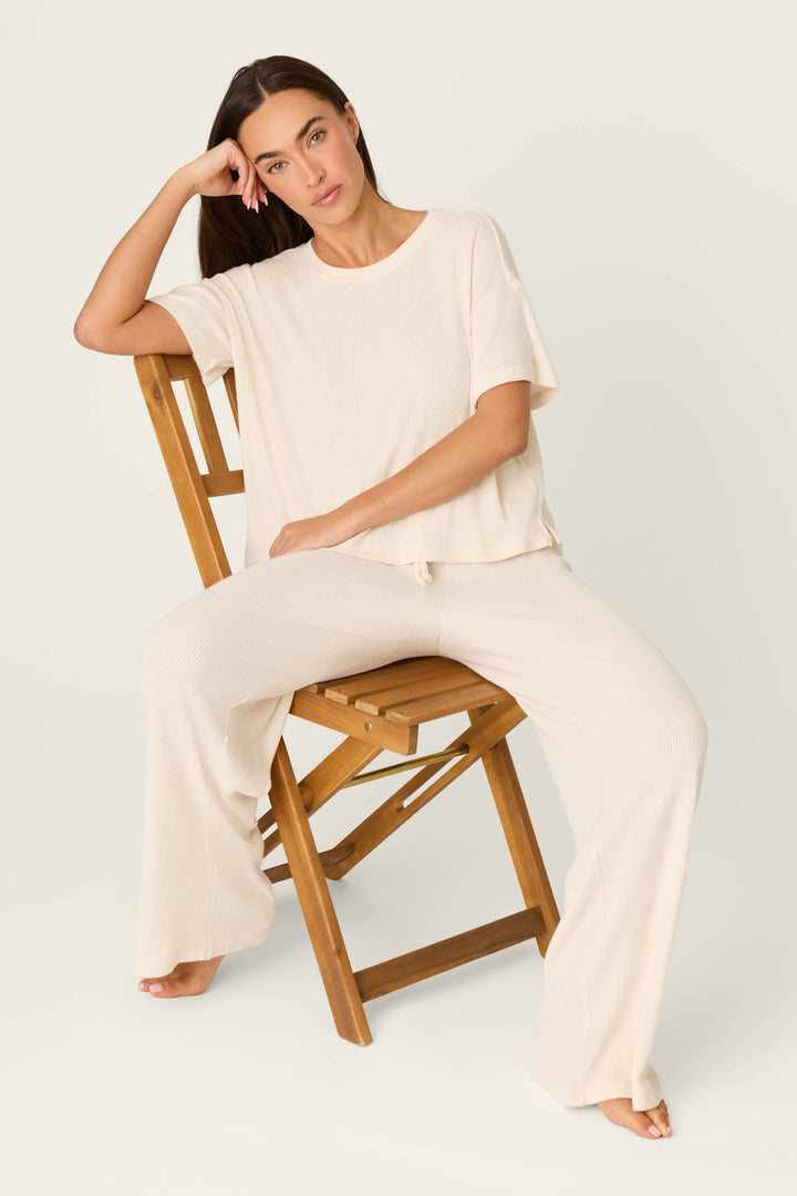 A woman with long brown hair sits barefoot on a wooden chair, wearing the P.J. Salvage Textured Essentials Stone Long PJ Set—a light beige, loose-fitting loungewear ensemble with a short-sleeve top and wide-leg pants—looking relaxed at the camera.