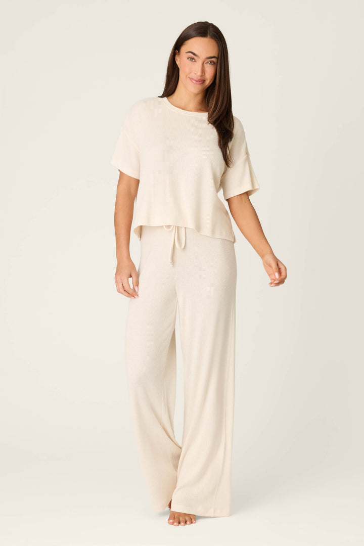 A woman stands barefoot against a plain background, wearing the P.J. Salvage Textured Essentials Stone Long PJ Set—a cozy light beige loungewear set with a loose short-sleeve top and wide-leg drawstring pants. She smiles softly at the camera.