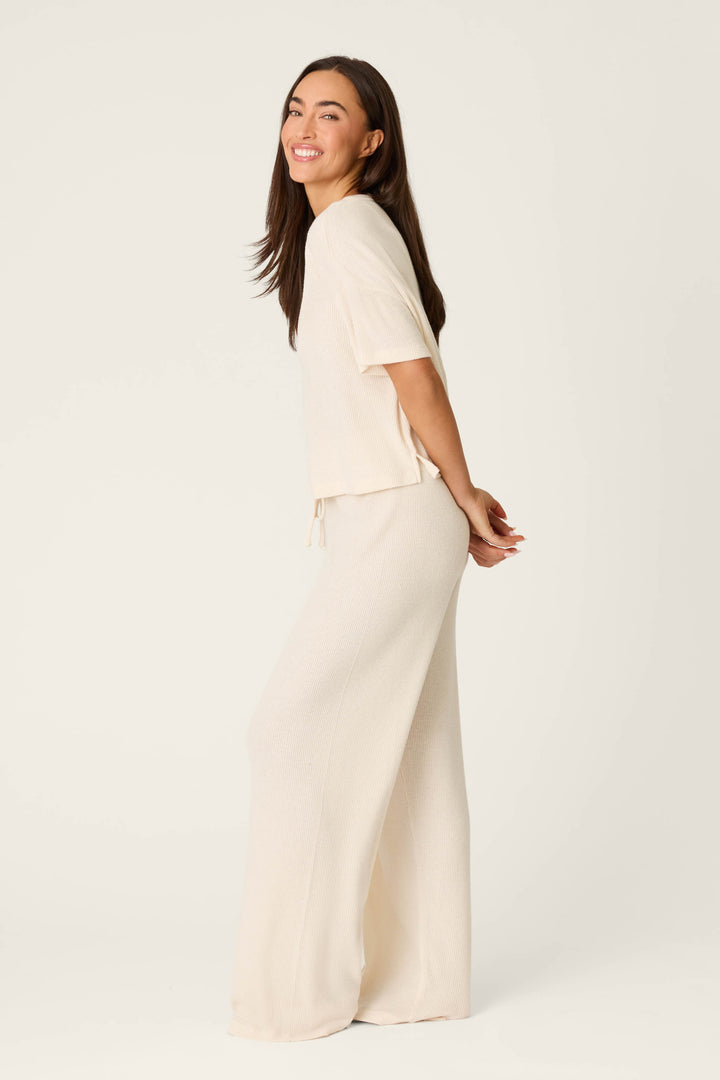 A woman with long brown hair smiles sideways, arms behind her back, modeling the P.J. Salvage Textured Essentials Stone Long PJ Set—an off-white short-sleeve top and wide-leg pants—against a plain light background.