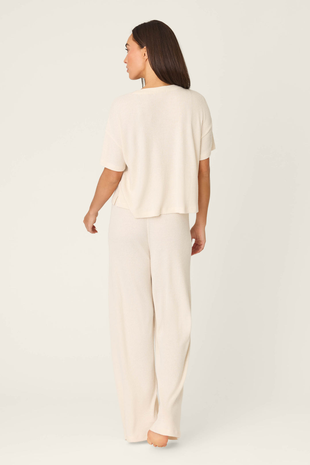 A person with long brown hair stands barefoot, facing away, wearing the P.J. Salvage Textured Essentials Stone Long PJ Set—a cozy rib knit loungewear set with a loose cream short-sleeve top and matching wide-leg pants—against a plain background.