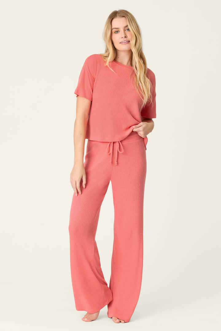 A person stands barefoot against a plain light background, wearing the P.J. Salvage Textured Essentials Sunset Rose Long PJ Set—a comfy coral pink short-sleeve top and drawstring wide-leg pants. Their long blonde hair is worn loose.