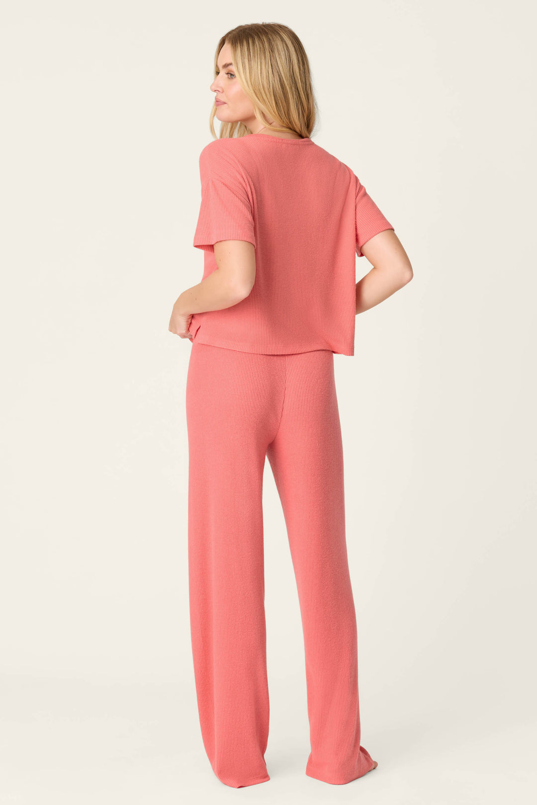 A woman stands facing away, head turned slightly, wearing the P.J. Salvage Textured Essentials Sunset Rose Long PJ Set—a comfy pink short-sleeve top with matching wide-leg pants—against a plain, light background.