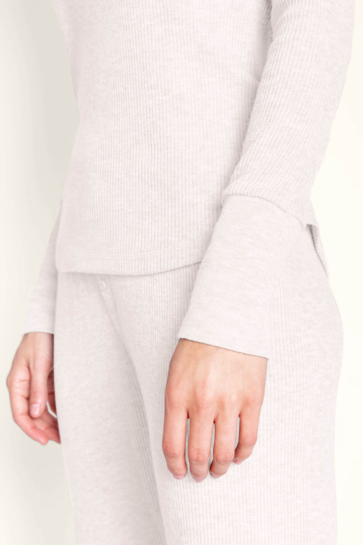 A person wearing the P.J. Salvage Textured Essentials Stone Lounge Set, featuring a light beige ribbed long-sleeve top and matching pants, stands with their left arm relaxed by their side. Only the torso, hips, and part of the left hand are visible against a neutral background.