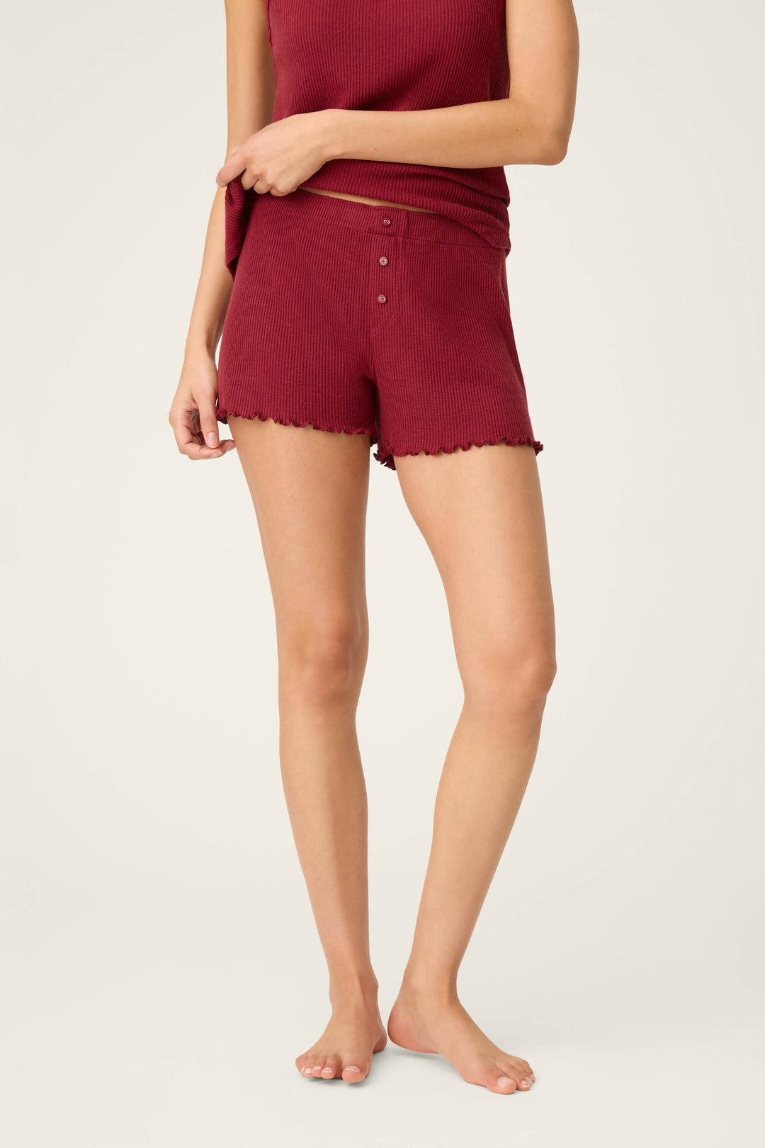 A woman models P.J. Salvage's Textured Essentials Port Shorts, featuring a dark red ribbed fabric, elastic waistband, lettuce hem, and button detail, paired barefoot with a matching tank top against a light backdrop.