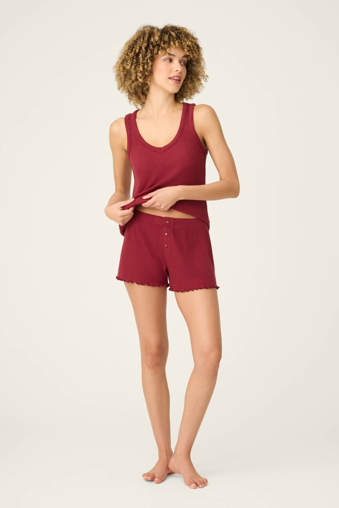 A woman with curly hair wears P.J. Salvage Textured Essentials Port Shorts and a matching sleeveless top, standing barefoot on a light background. She holds the hem of her top and looks slightly to the side.