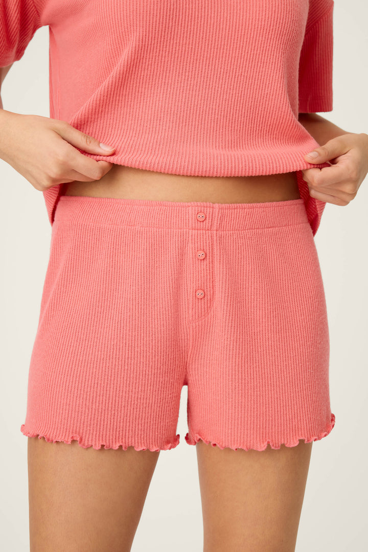 A person wears the P.J. Salvage Textured Essentials Sunset Rose Shorts, part of a matching pink knit set with a short-sleeve top and high-waisted shorts featuring three buttons and ruffled hems, holding up the hem of the top.