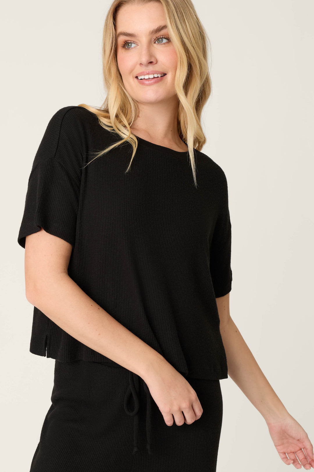 A person with long blonde hair wears the P.J. Salvage Textured Essentials Black Short Sleeve Top and a matching drawstring skirt, standing against a plain light background and smiling slightly.