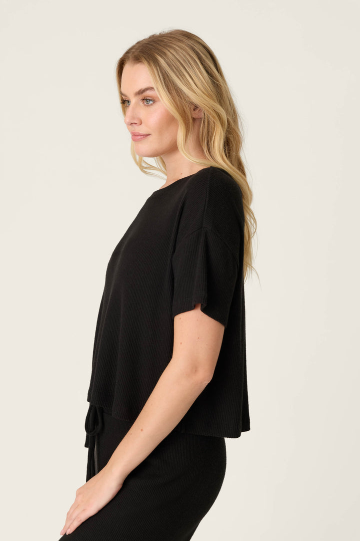 A woman with long blonde hair stands in profile against a plain background, wearing the P.J. Salvage Textured Essentials Black Short Sleeve Top and black bottoms.