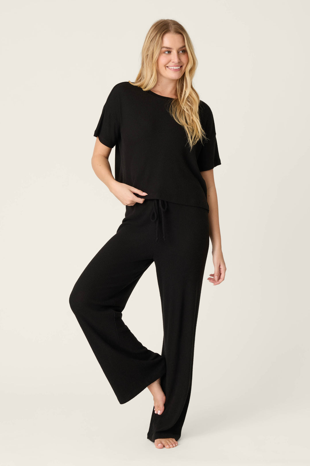 A person with long blonde hair smiles barefoot on one leg against a plain light background, wearing the P.J. Salvage Textured Essentials Black Short Sleeve Top with matching wide-leg pants.