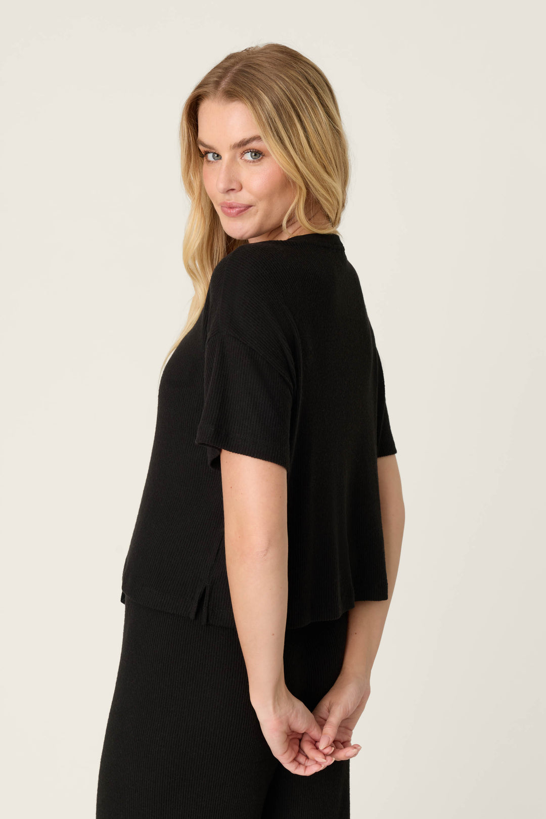 A person with long blonde hair models the P.J. Salvage Textured Essentials Black Short Sleeve Top, paired with a matching black skirt, standing against a plain light background and facing partially away from the camera.