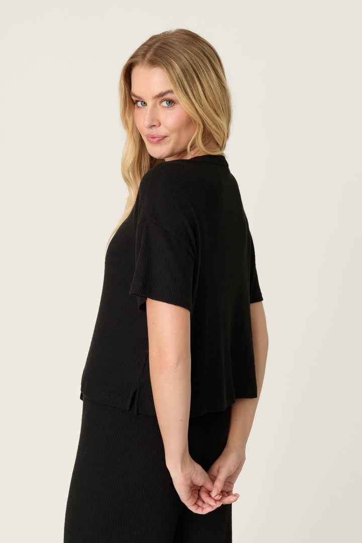 A person with long blonde hair models the P.J. Salvage Textured Essentials Black Short Sleeve Top, paired with a matching black skirt, standing against a plain light background and facing partially away from the camera.