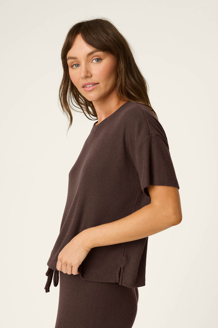 A woman with long brown hair is wearing the P.J. Salvage Textured Essentials Chocolate Brown Short Sleeve Top, paired with matching pants, standing sideways and looking at the camera against a plain light background.