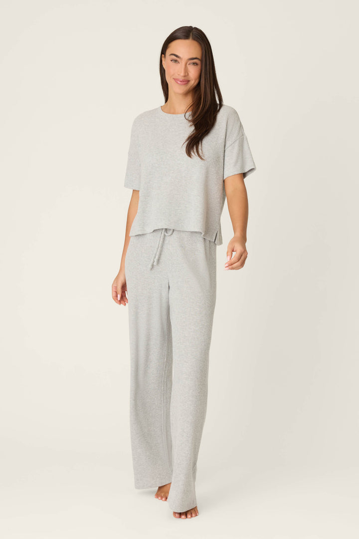 A woman with long brown hair stands barefoot against a plain background, wearing the P.J. Salvage Textured Essentials Heather Grey Short Sleeve Top and matching wide-leg drawstring pants, with a neutral expression.