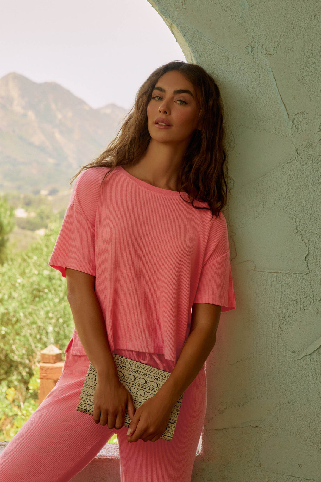 A woman with long brown hair wears the P.J. Salvage Textured Essentials Sunset Rose Short Sleeve Top and matching pants, standing against a light green textured wall with a beige patterned clutch, mountains, and lush greenery in the background.