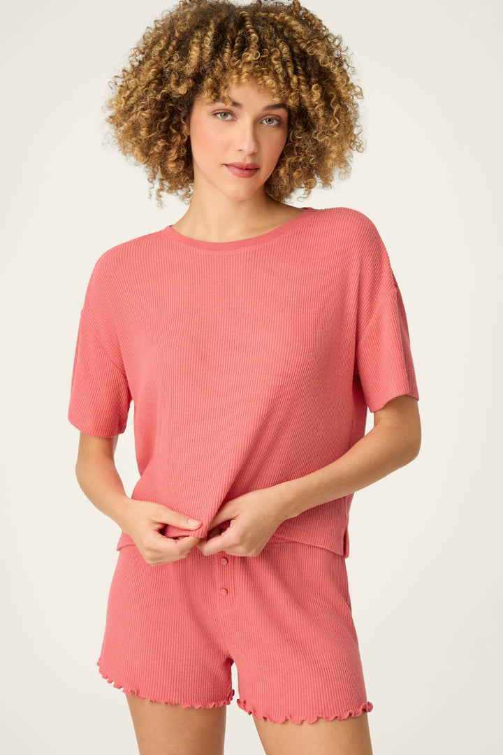 A person with curly hair wears the P.J. Salvage Textured Essentials Sunset Rose Short Sleeve Top and matching shorts, standing against a plain light background and holding the hem of the top with both hands.