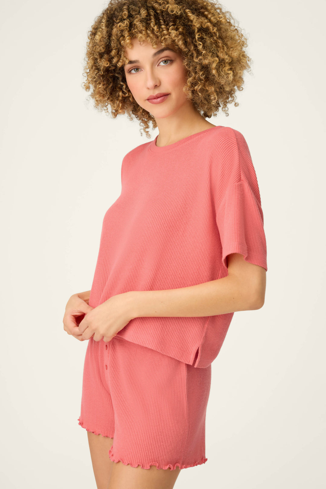 A person with curly hair is wearing the P.J. Salvage Textured Essentials Sunset Rose Short Sleeve Top and matching pink shorts, looking comfortable while standing against a plain light background.