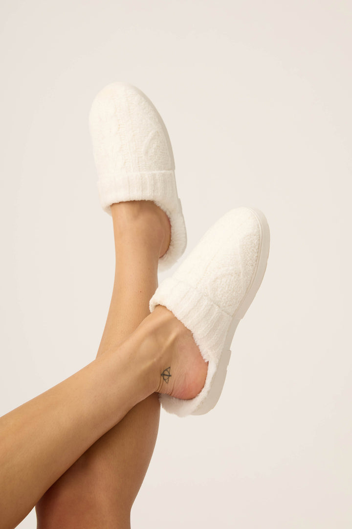 A person with crossed legs wears P.J. Salvages SLIDE CABLE CREW LOUNGE cozy white slippers, revealing a small ankle tattoo against a plain light background.