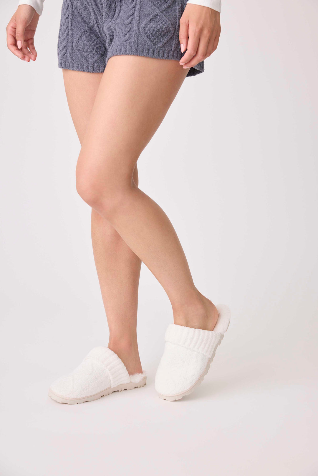 A person wearing gray knit shorts and white P.J. Salvage Chenille Cozy Cable Slippers stands with one leg crossed in front of the other against a plain light background.