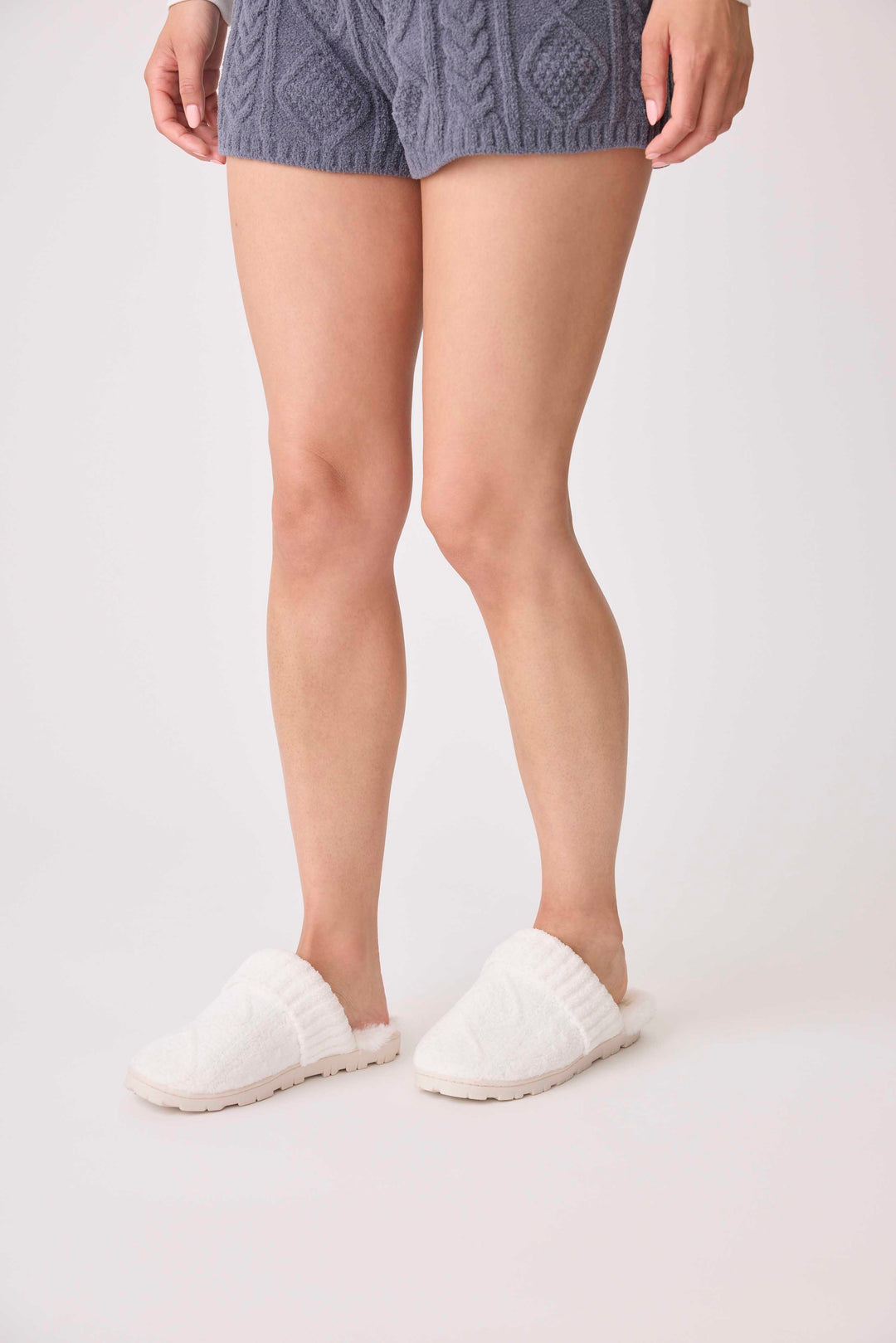 A person is shown from the waist down on a white surface, wearing gray knit shorts and P.J. Salvage Chenille Cozy Cable Slippers in white.