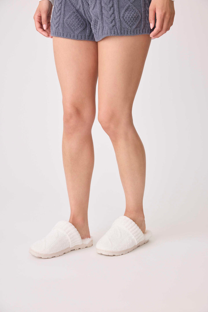 A person is shown from the waist down on a white surface, wearing gray knit shorts and P.J. Salvage Chenille Cozy Cable Slippers in white.