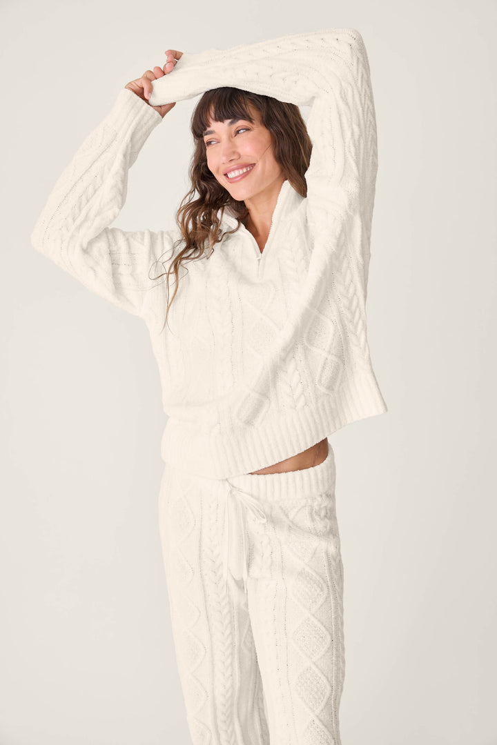 A person smiles and lifts one arm above their head while wearing the PJ SET CABLE CREW LOUNGE by P.J. Salvage— a matching white cable-knit sweater and pants— against a plain, light background.