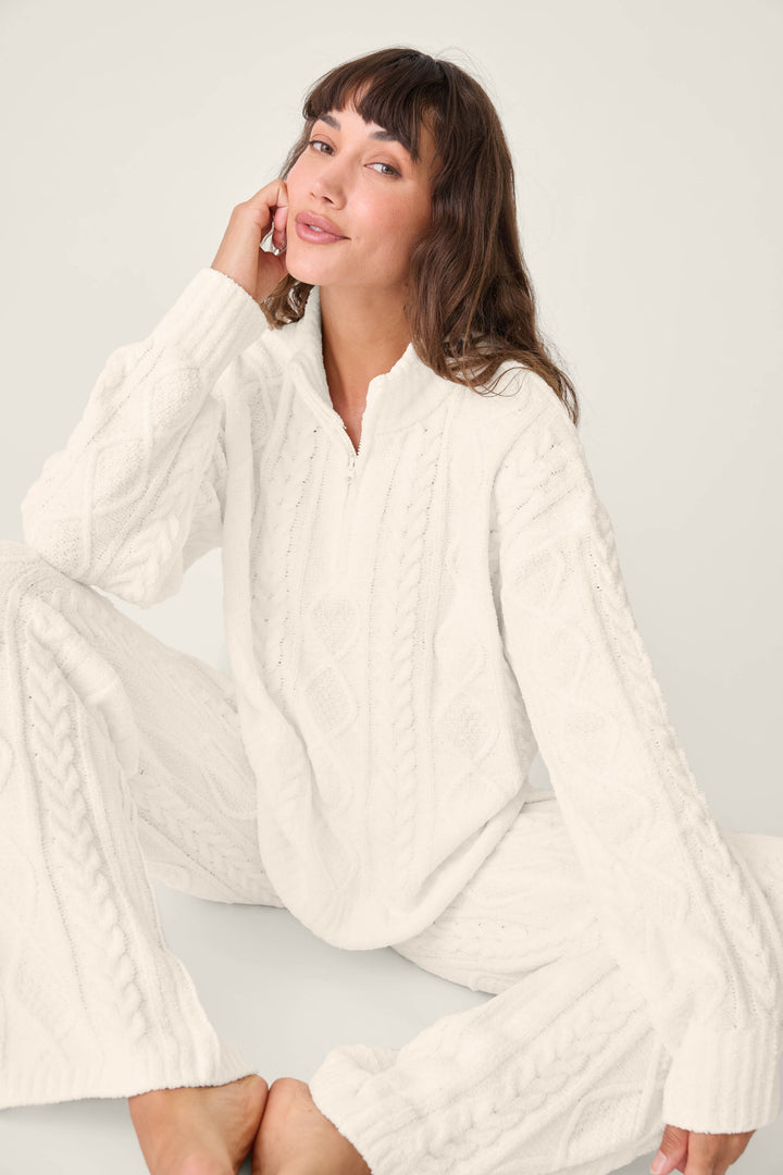 A woman with brown hair and bangs sits barefoot on the floor, wearing the PJ SET CABLE CREW LOUNGE by P.J. Salvage—a matching white cable-knit sweater and pants set—looking at the camera against a plain background.