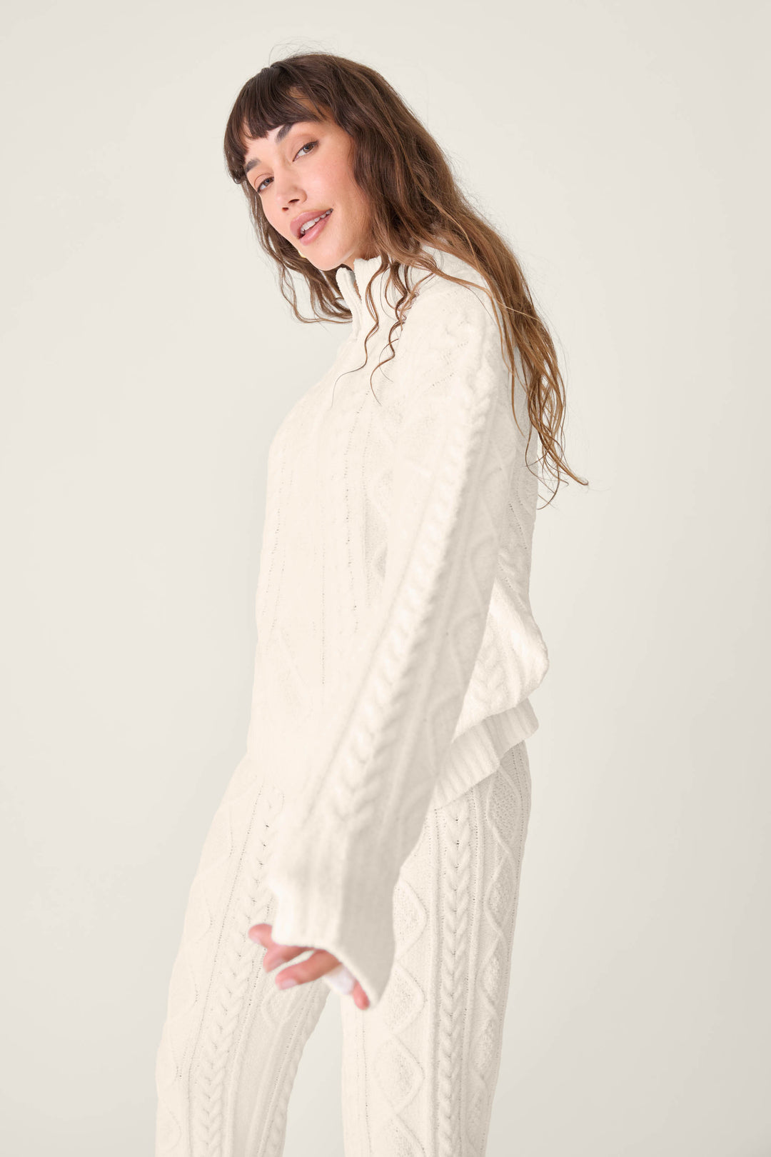 A person with long brown hair poses in the P.J. Salvage PJ SET CABLE CREW LOUNGE—matching white cable-knit sweater and pants—against a plain light background.