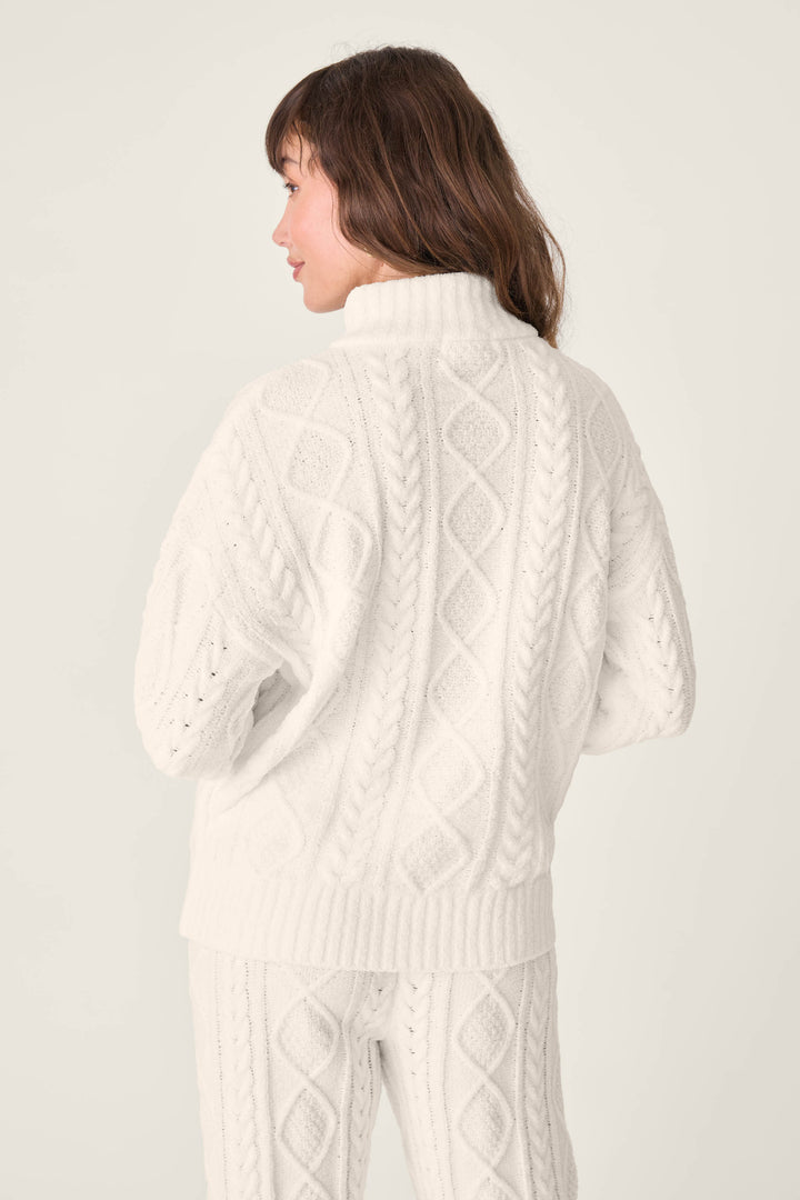 A woman is seen from behind wearing the PJ SET CABLE CREW LOUNGE by P.J. Salvage—a white, high-collar cable-knit sweater with diamond and braid patterns, paired with matching pants. The background is simple and light-colored.