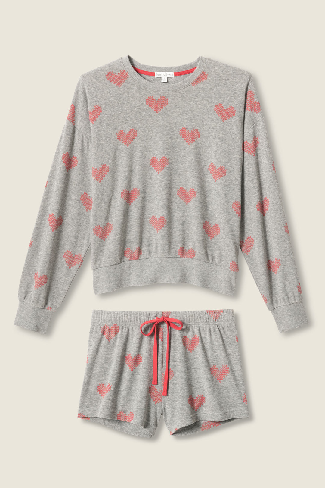 The P.J. Salvage Heart Print Fleece PJ Set features a gray long-sleeve top and drawstring shorts with an all-over pink heart pattern and a pink waistband tie, styled flat on a light background.