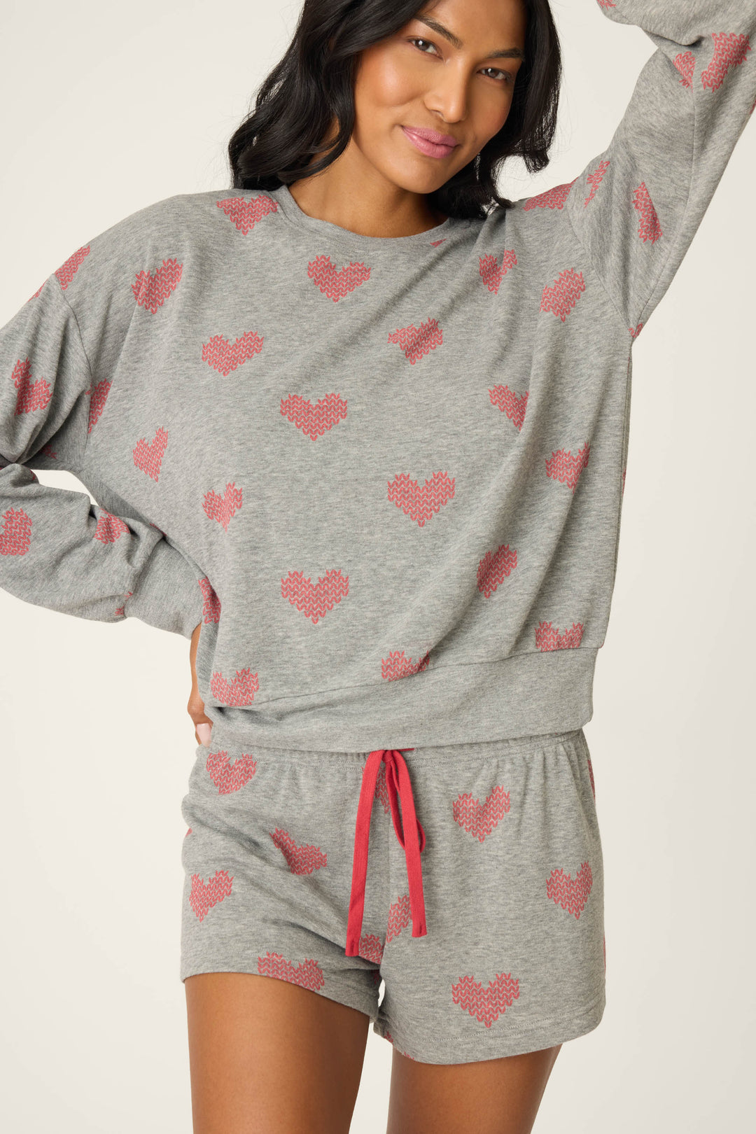 A person wearing the P.J. Salvage Heart Print Fleece PJ Set, featuring a matching grey long-sleeve top and shorts with red hearts and a red drawstring, stands with one hand on their hip and the other arm raised.