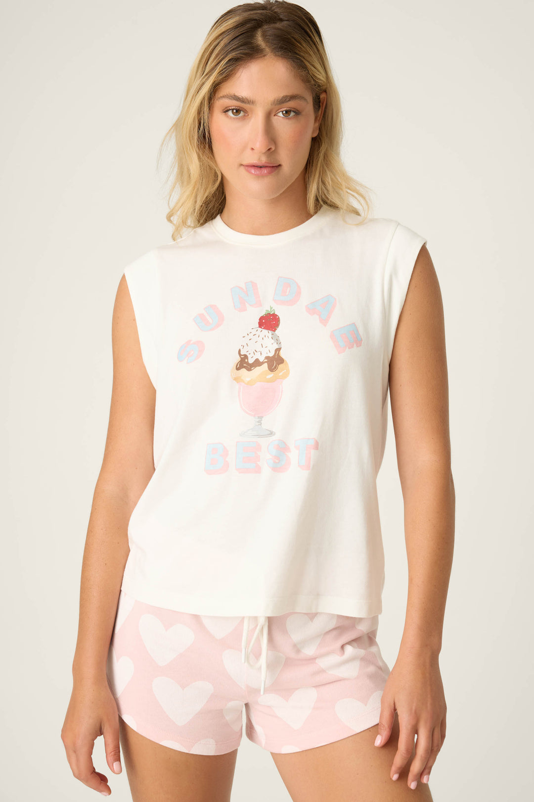 Model wearing ivory muscle tee with ‘Sundae Best’ graphic and pink shorts featuring white heart print.
