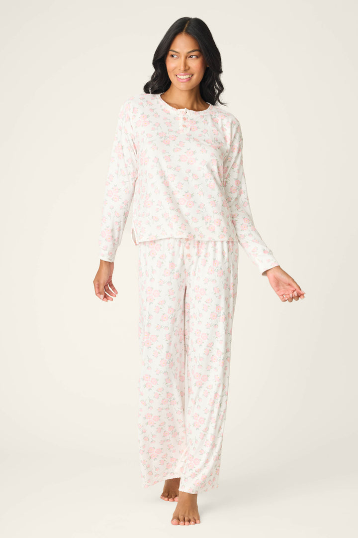 A woman stands barefoot, smiling to the side in the P.J. Salvage Ivory Floral Print Henley PJ Set—a matching long-sleeve pajama set with a light pink floral pattern on a white background, one arm slightly raised.