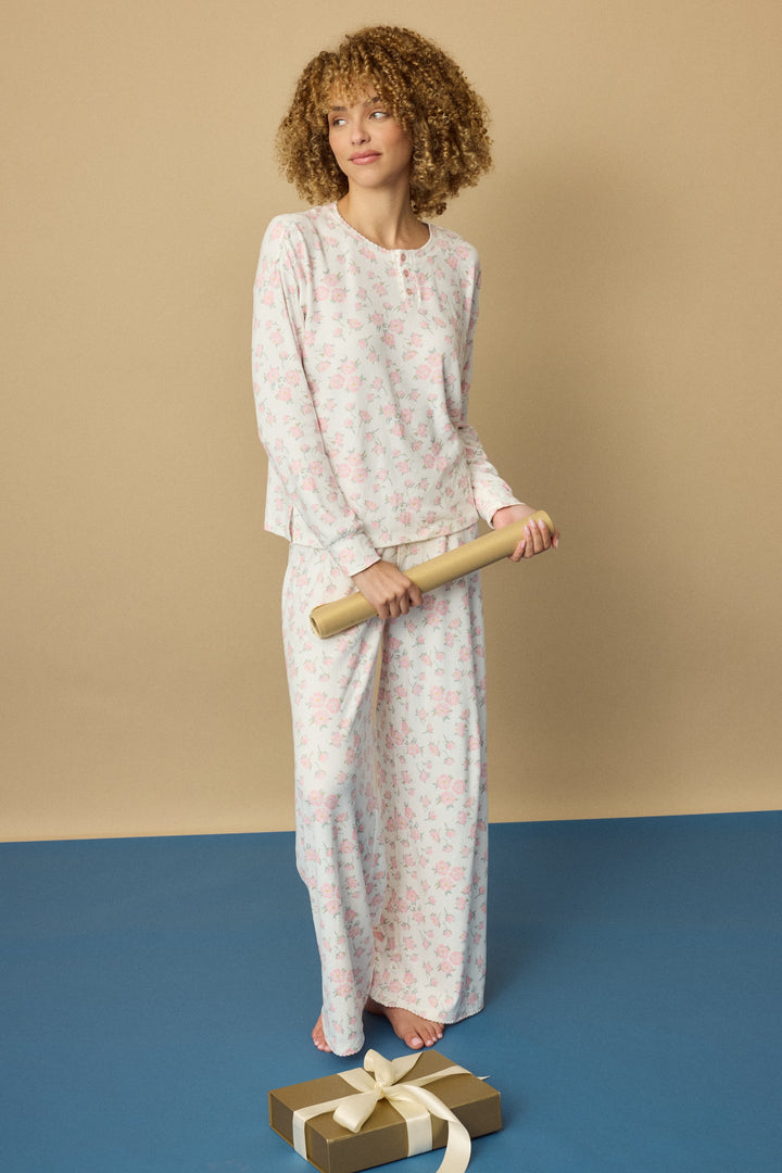 A person with curly hair stands barefoot on a blue floor wearing the P.J. Salvage Ivory Floral Print Henley PJ Set, holding a rolled-up paper. A gold-wrapped gift with a white ribbon is placed in front of them.
