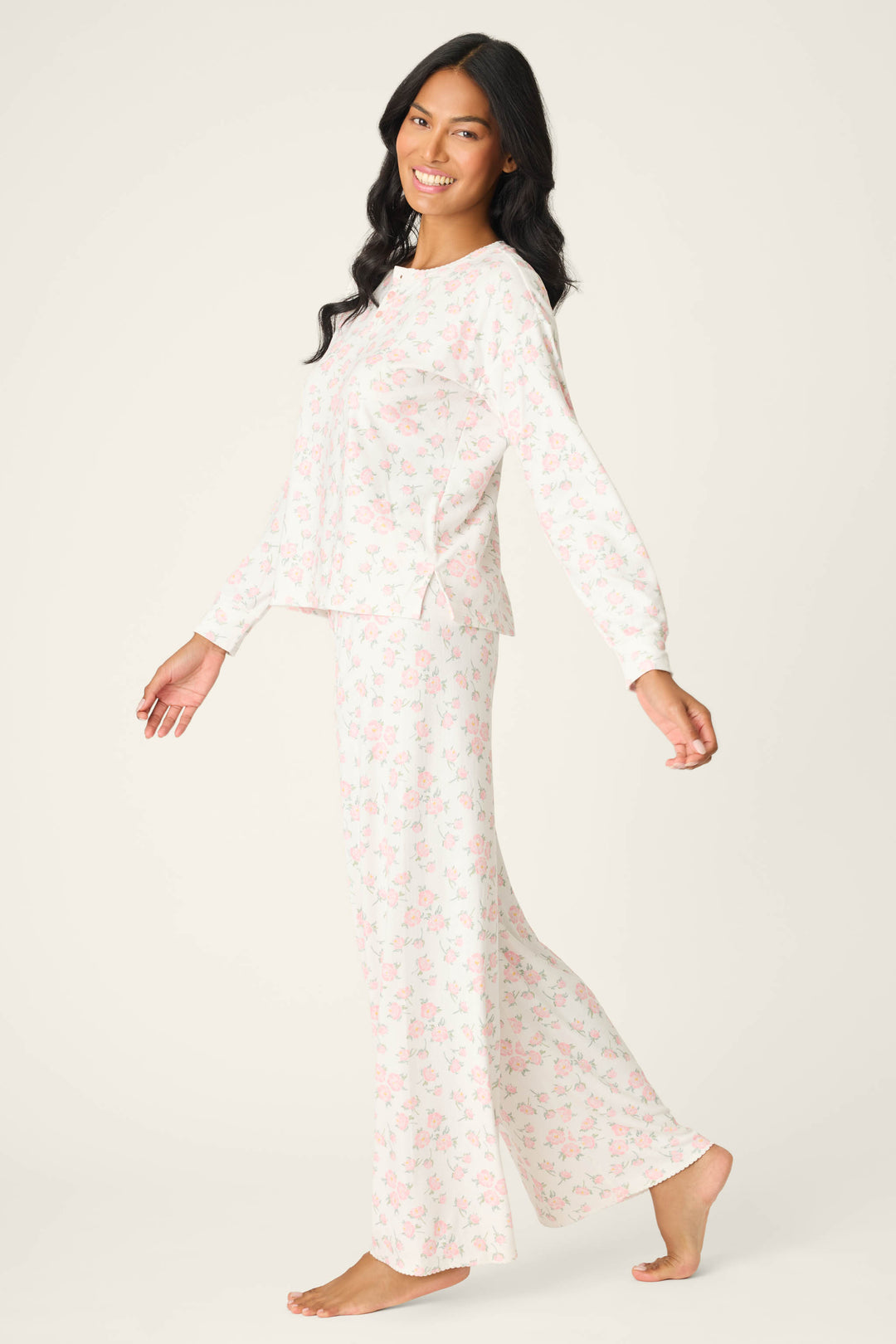 A woman with long dark hair wears the P.J. Salvage Ivory Floral Print Henley PJ Set, featuring light-colored, loose-fitting pajamas with a pink floral pattern. She stands barefoot and smiles against a plain light background.