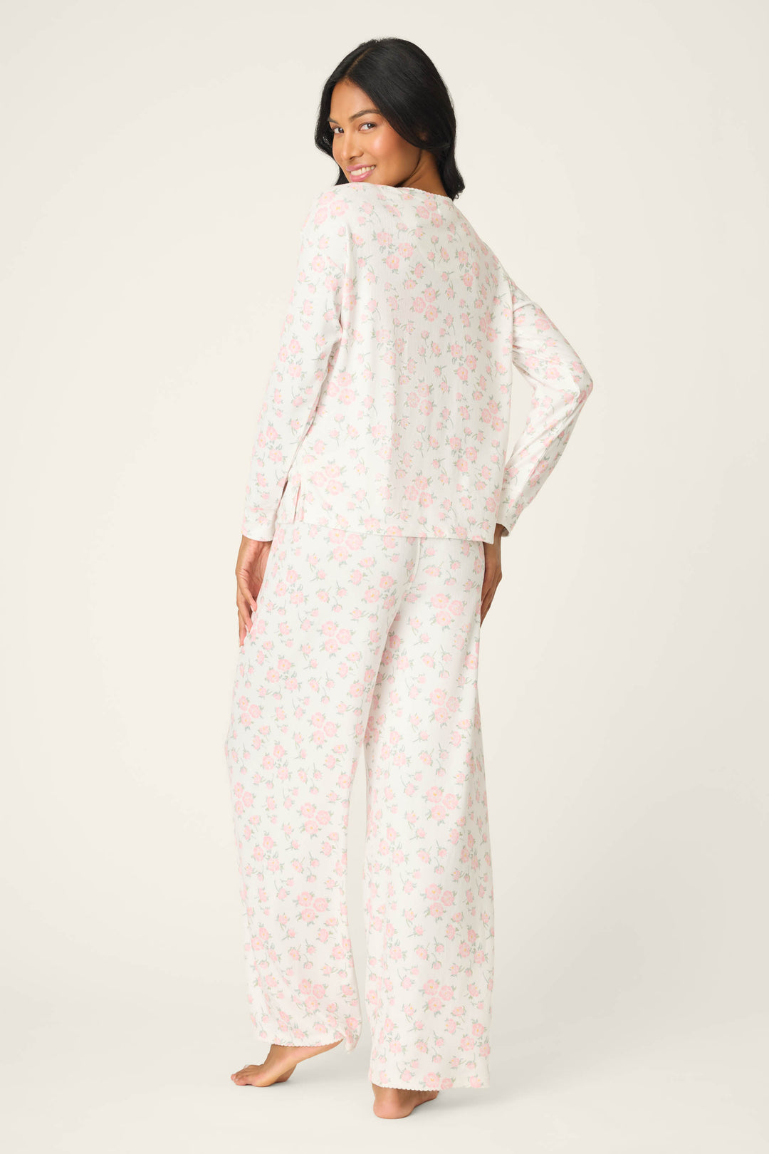 A woman poses barefoot in the P.J. Salvage Ivory Floral Print Henley PJ Set, featuring a long-sleeve top and loose pants with light pink and white floral design, facing away from the camera and glancing over her shoulder.