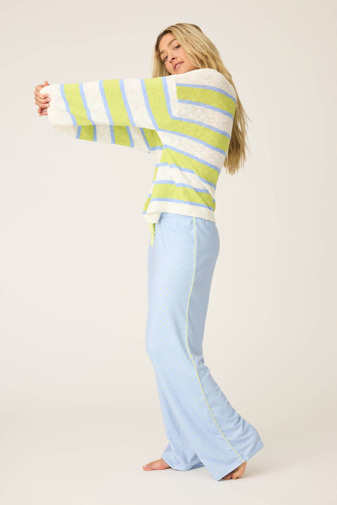 A woman stands barefoot on a light background, stretching her arms out while wearing P.J. Salvage’s Beautiful Life Striped Snuggle Sweater—featuring green, white, and gray stripes—paired with light blue pajama pants.