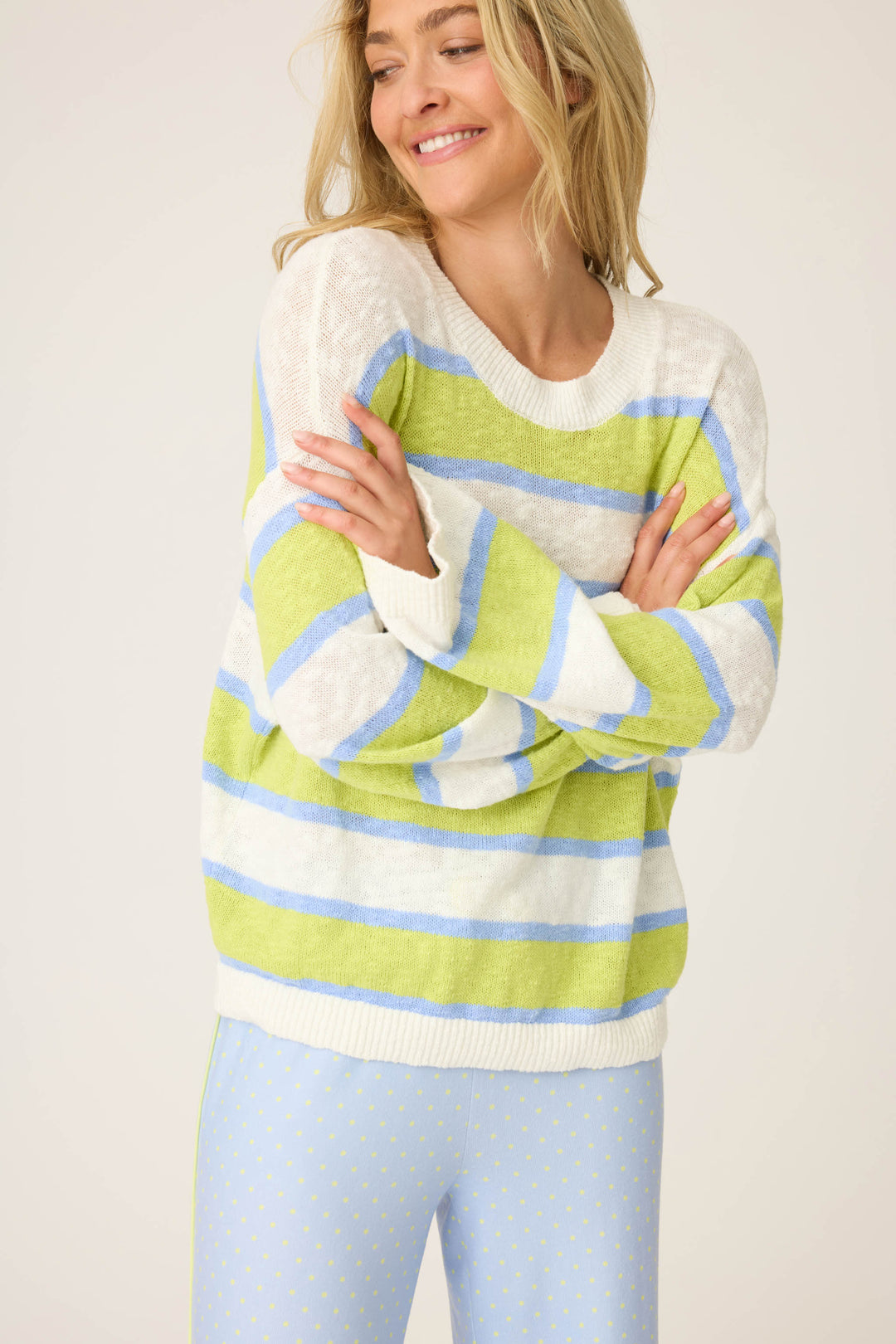A woman with long blonde hair smiles with arms crossed, wearing the P.J. Salvage Beautiful Life Striped Snuggle Sweater—white with green and blue stripes—paired with pale blue pants featuring tiny yellow dots. The background is plain and light.