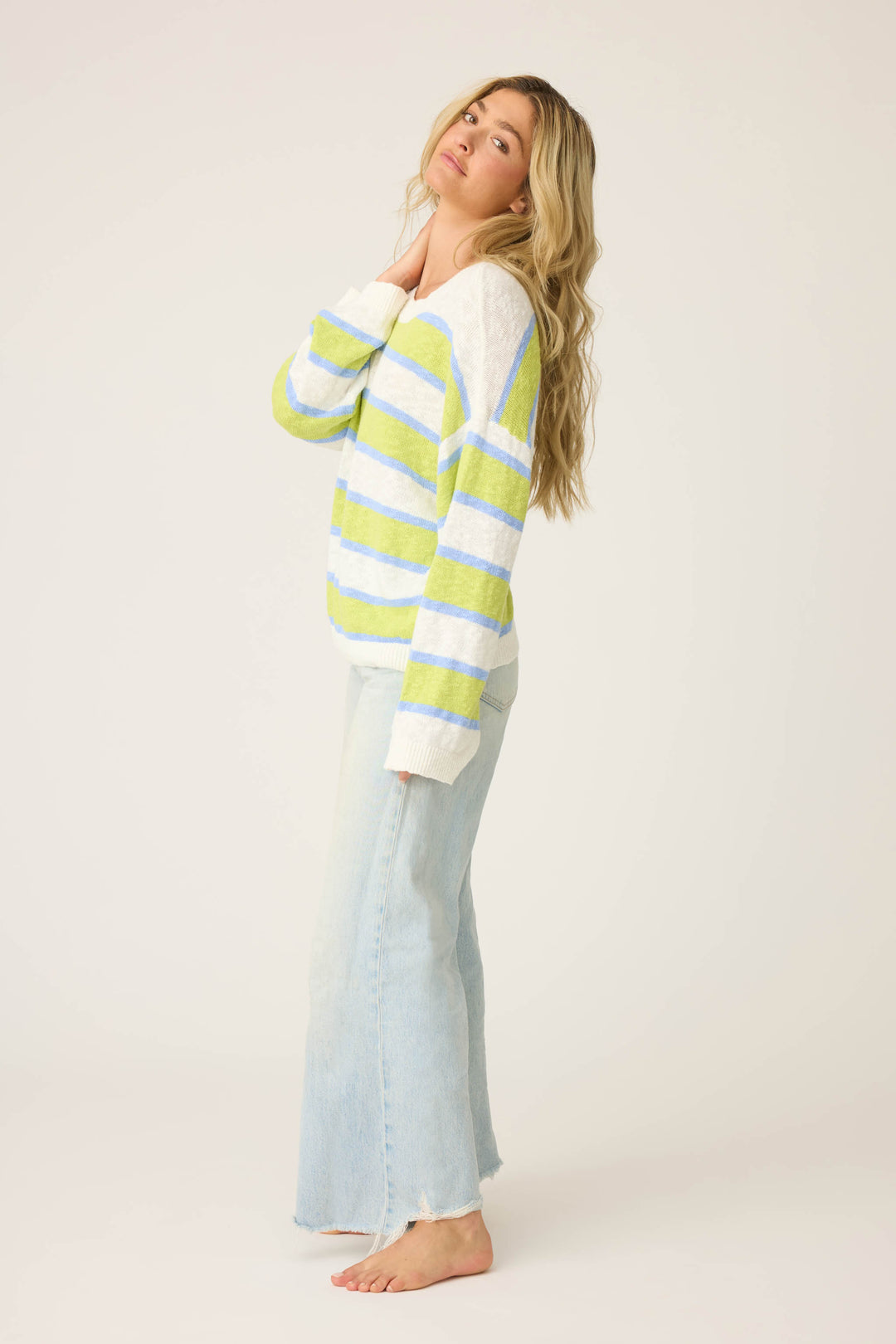A person with long, wavy blonde hair models the P.J. Salvage Beautiful Life Striped Snuggle Sweater—green, white, and blue stripes—paired with light blue jeans, barefoot, posing against a plain light background.