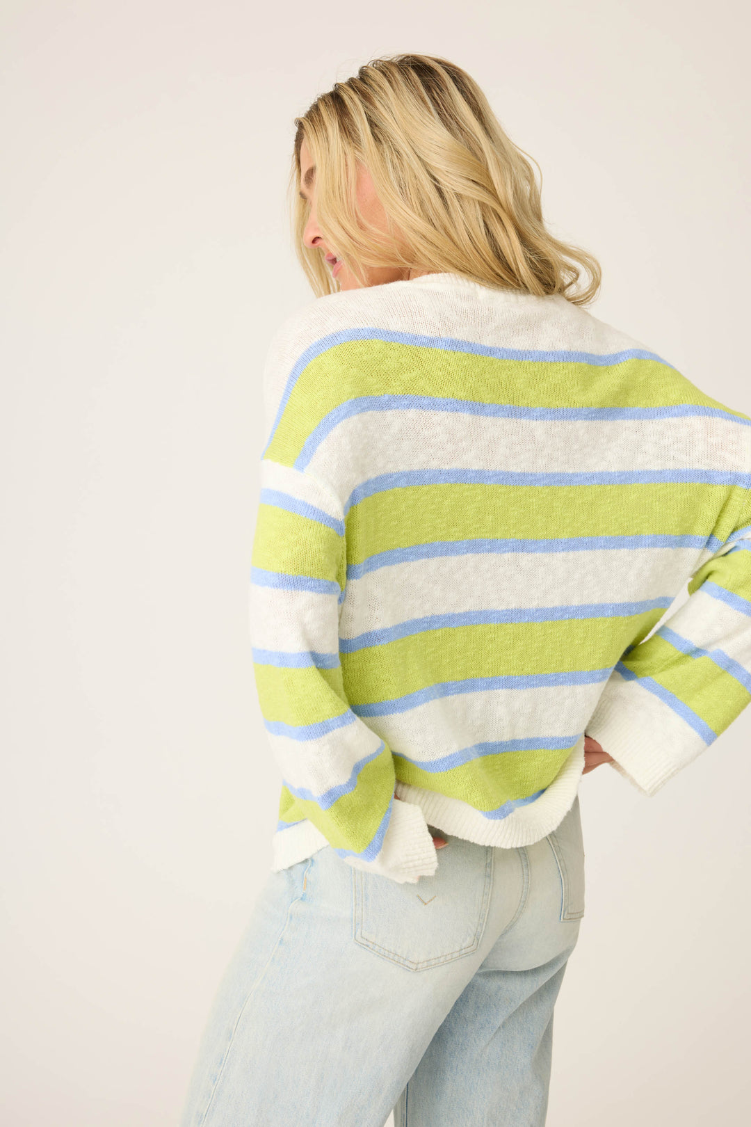 A person with blonde hair, facing away from the camera, wears the P.J. Salvage Beautiful Life Striped Snuggle Sweater in white, light blue, and light green stripes, paired with light blue jeans against a plain background.