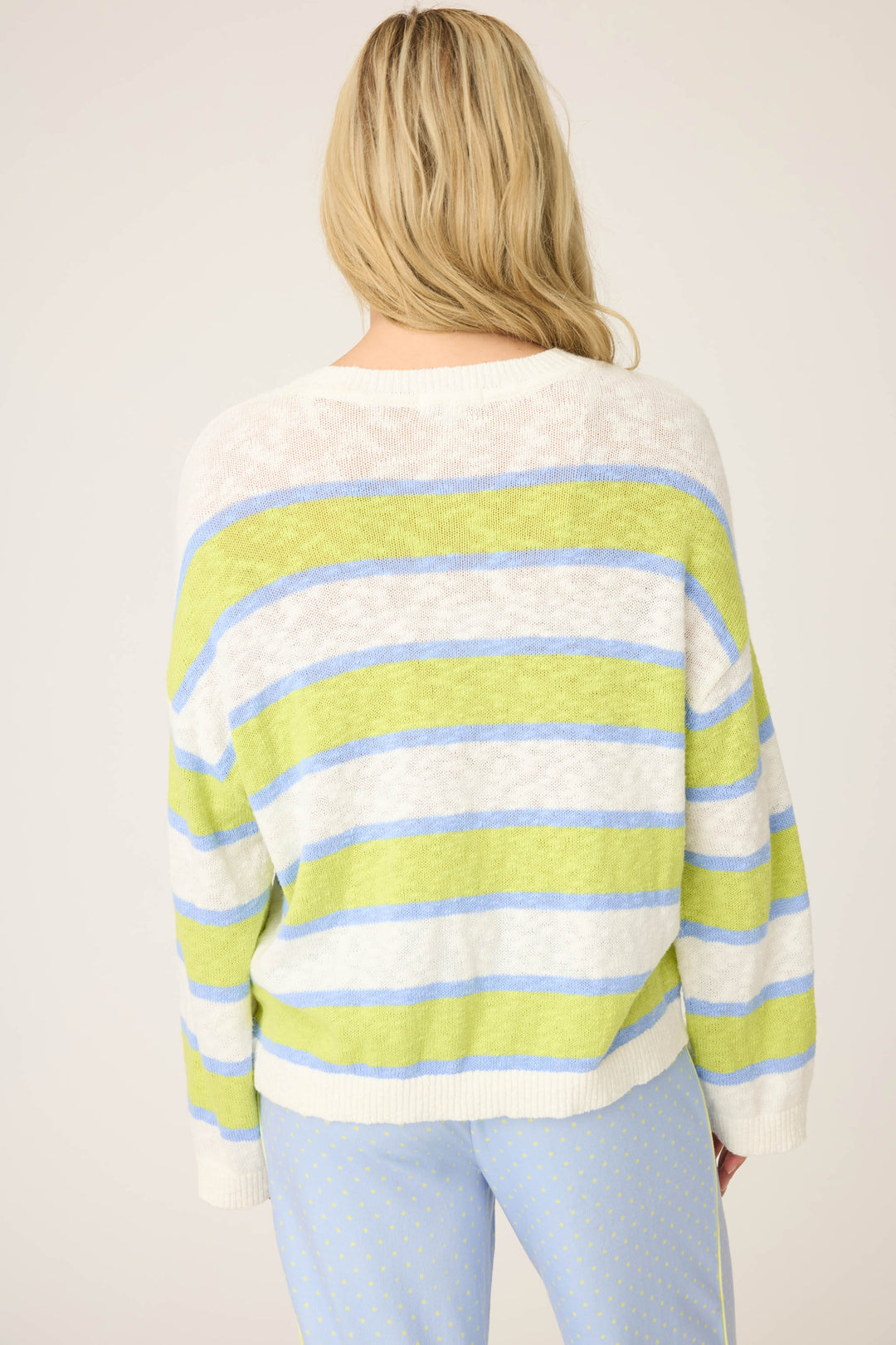 A person with long blonde hair is seen from behind wearing the P.J. Salvage Beautiful Life Striped Snuggle Sweater, featuring green, blue, and white stripes, paired with light blue dotted pants against a plain, light background.